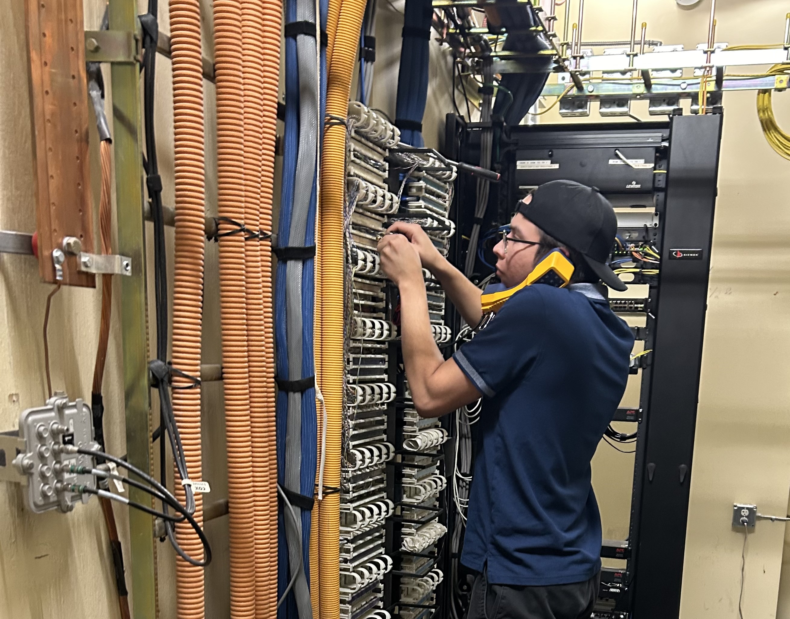 Picture of a technician standing in a telecommunications room testing phone lines with a phone to his ear.